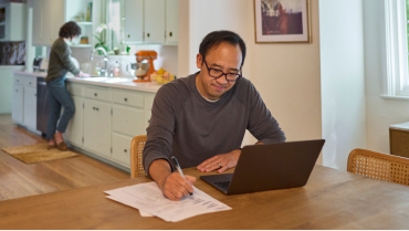 Man sitting at a kitchen table looking at a laptop and filling out paper forms next to him. A woman is in background at the kitchen sink.