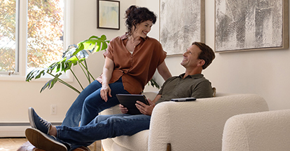 Man sitting in a chair with his feet propped up while holding a digital tablet. A woman is sitting on the arm of the chair and looking down at him and smiling.